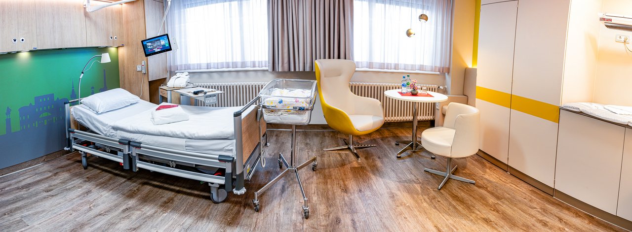 Panoramaansicht eines Krankenhauszimmers. Links ein Krankenbett vor grüner Wand. Rechts vor dem Fenster eine Sitzgruppe, am rechten Bildrand eine Schrankwand mit Wickeltisch.