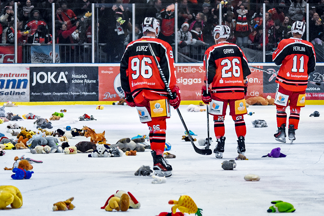 Drei Eishockey-Spieler der Saale Bulls sind von hinten auf dem Eis zu sehen. Auf dem Eis liegen überall verteilt viele Kuscheltiere.