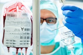 Prof. Ulrich Ronellenfitsch steht vor einem Ständer mit einer Bluttransfusion und blickt in die Kamera. In seiner linken Hand hält er ein Glasfläschchen mit einer klaren Flüssigkeit und der Beschriftung "1g Tranexamsäure".