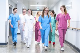 Eine Gruppe junger Menschen in medizinischer Arbeitskleidung in verschiedenen Farben läuft miteinander redend einen Gang entlang.