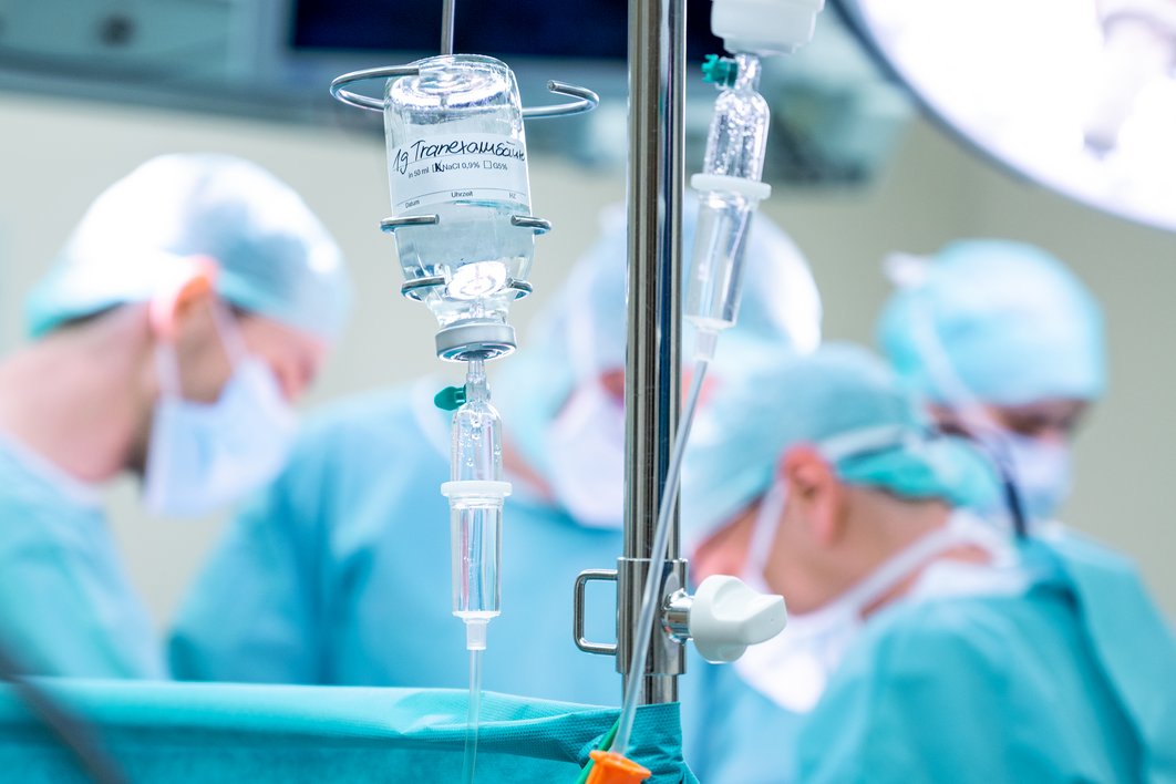 Im Vordergrund ist ein Glasfläschchen mit einer klaren Flüssigkeit und der Aufschrift "1g Tranexamsäure" zu erkennen. Im Hintergrund sieht man vier Personen, die im Operationssaal arbeiten.
