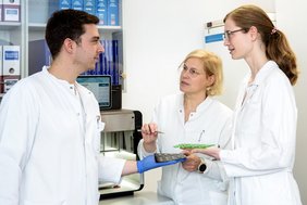 Drei Personen in medizinischer Arbeitskleidung stehen an einem 3D-Drucker und halten gedruckte Medikamente in den Händen.