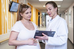 Eine Frau im weißen Kittel hält ein Tablet und ist mit einer Schwangeren mit weißem Oberteil im Gespräch. Beide stehe im Korridor einer Klinik.