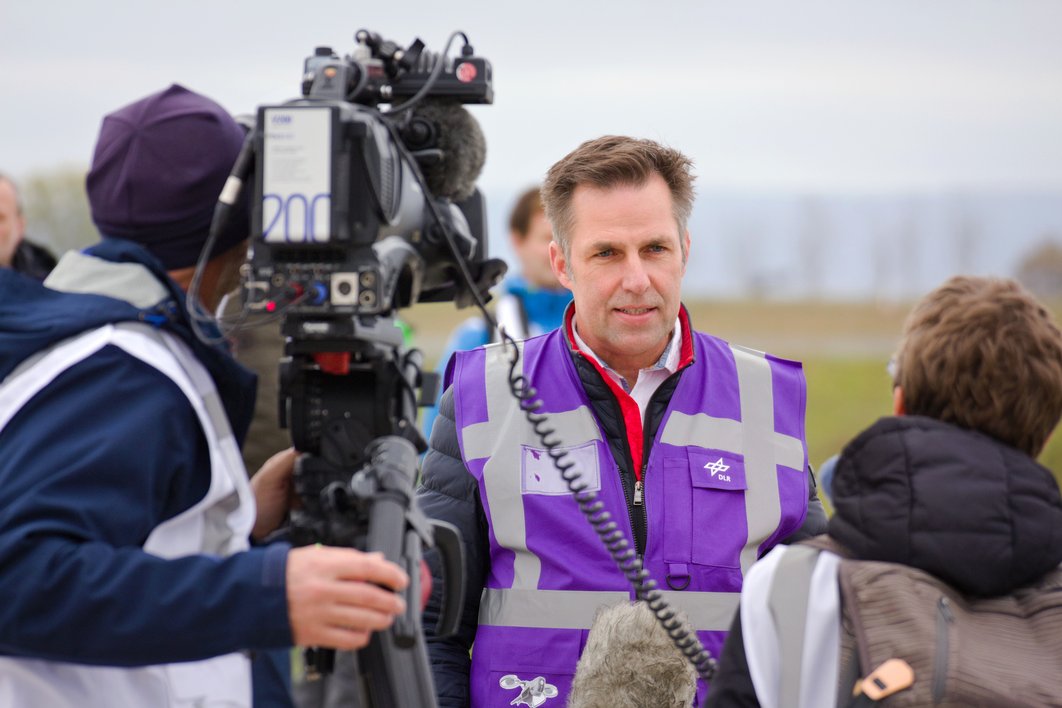 Ein Mann in lila Weste wird von einem Kameramann und einer Reporterin interviewt.