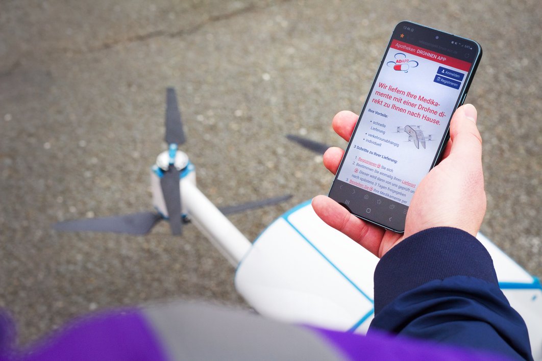 Eine Hand hält ein Handy mit der Apotheken-Drohnen-App auf dem Display. Auf dem Boden ist die Drohne zu erkennen.