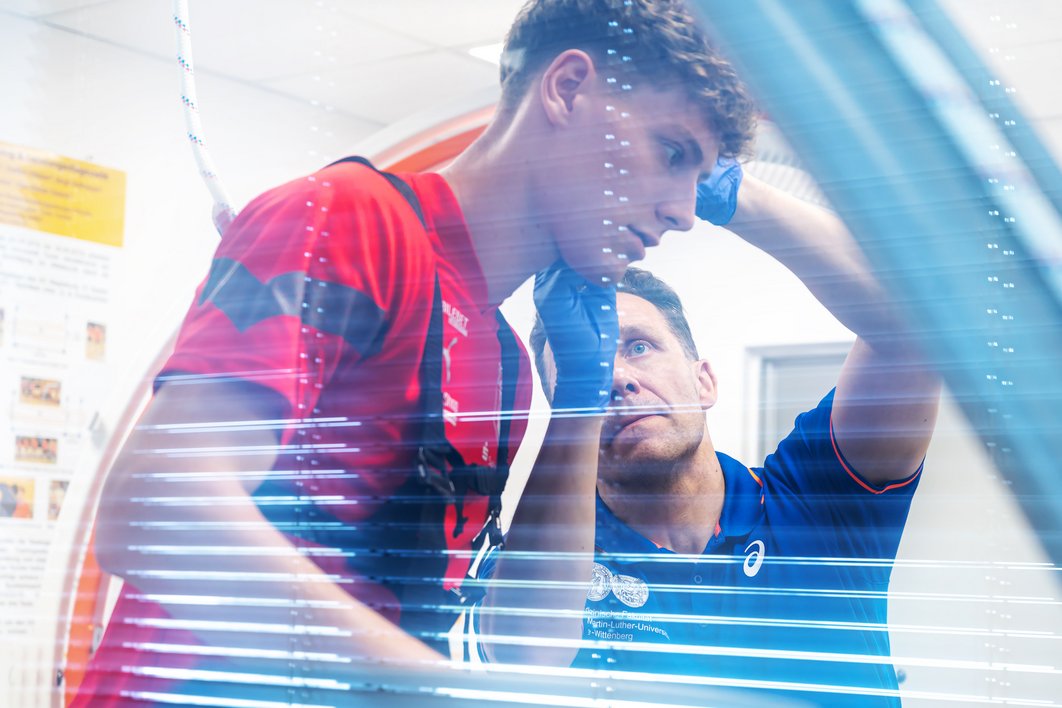 Zwei Männer sind durch ein Fenster zu sehen. Der Mann rechts mit blauem Shirt (Prof. Rene Schwesig) nimmt gerade Blut vom Ohr des Mannes, der links von ihm steht (Seymour Fünger) und ein rotes HFC-Trikot trägt
