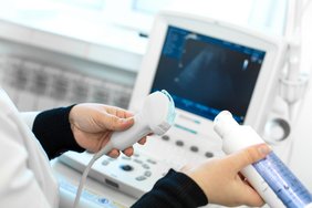 Es sind zwei Hände zu sehen, die Gleitmittel auf den Schallkopf eines Sonografie-Geräts auftragen. Im Hintergrund ist unscharf ein Ultraschallmonitor zu erkennen. 