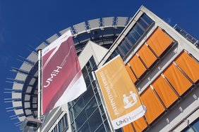 Zwei Flaggen vorm Hauptgebäude des Universitätsklinikums Halle (Saale). Die linke Fahne zeigt das Logo der Universitätsmedizin Halle auf rotem Grund, die rechte Fahne den Schriftzug "Patientensicherheit" auf Orange.