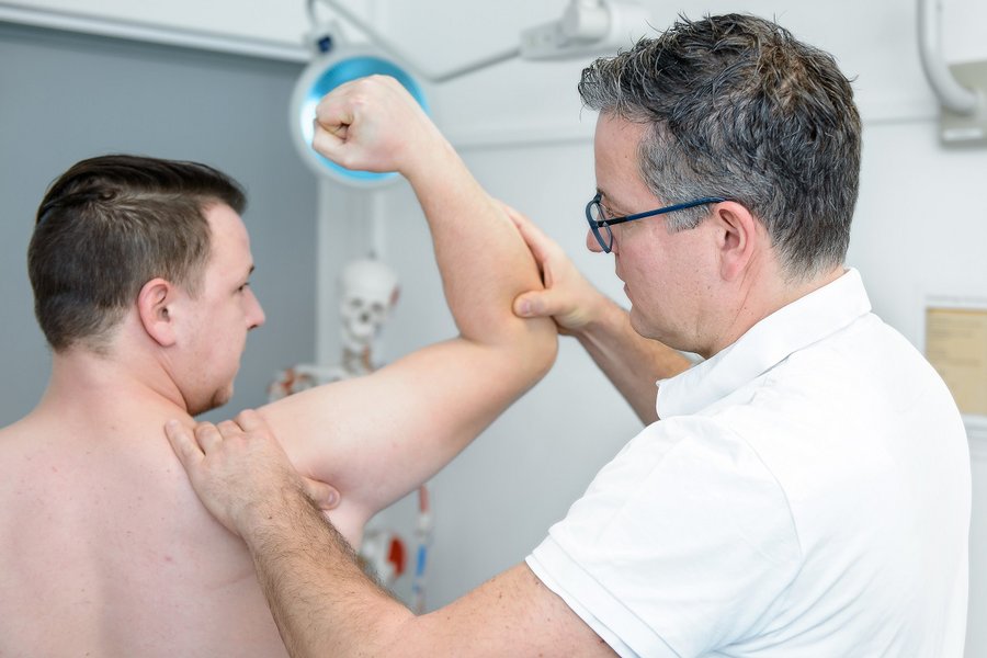 Ein Arzt (rechts im Bild) untersucht die Schulter eines Patienten.  Ein Arzt (rechts im Bild) untersucht die Schulter eines Patienten.