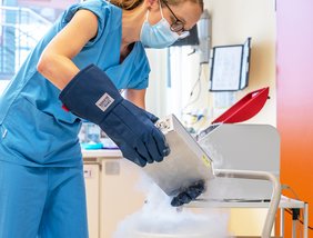 Eine Frau in medizinischer Arbeitskleidung trägt Handschuhe und hält eine silberne Kiste in den Händen. Sie befindet sich in einer medizinischen Arbeitsumgebung.