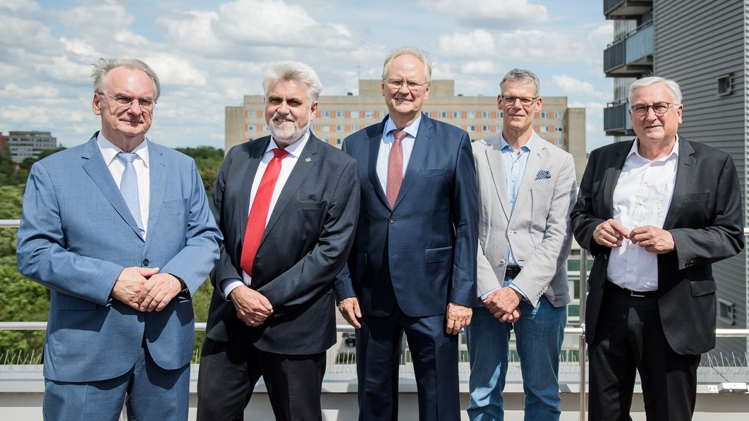 Reiner Haseloff, Prof. Dr. Armin Willingmann, Prof. Dr. Thomas Moesta, Egbert Geier und Michael Richter stehen nebeneinander auf einem Hausdach. Im Hintergrund sieht man das alte Bettenhaus des Universitätsklinikums Halle.