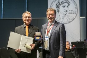Zwei Herren im Anzug halten eine Urkunde und eine Medaille lächelnd in die Kamera
