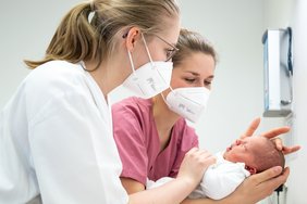 zwei Frauen in weißer und brombeerfarbener medizinischer Arbeitskleidung halten ein neugeborenes Baby