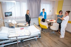 Krankenhauszimmer von schräg oben. Links ist ein Krankenbett zu sehen. im rechten oberen Bildrand stehen vier Frauen an einer gelben Sitzgruppe in einem Gespräch zusammen.