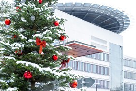 Im Vordergrund ist ein geschmückter und verschneiter Weihnachtsbaum zu sehen; im Hintergrund ist das Hauptgebäude des Universitätsklinikums Halle (Saale) zu erkennen.