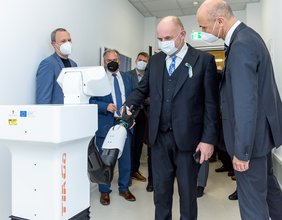 In einem Korridor stehen mehrere Personen mit Mund-Nasen-Schutz. Der Mann im Vordergrund schüttelt einem Roboter zur Medizinischen Versorgung die künstliche Hand und lächelt dabei.