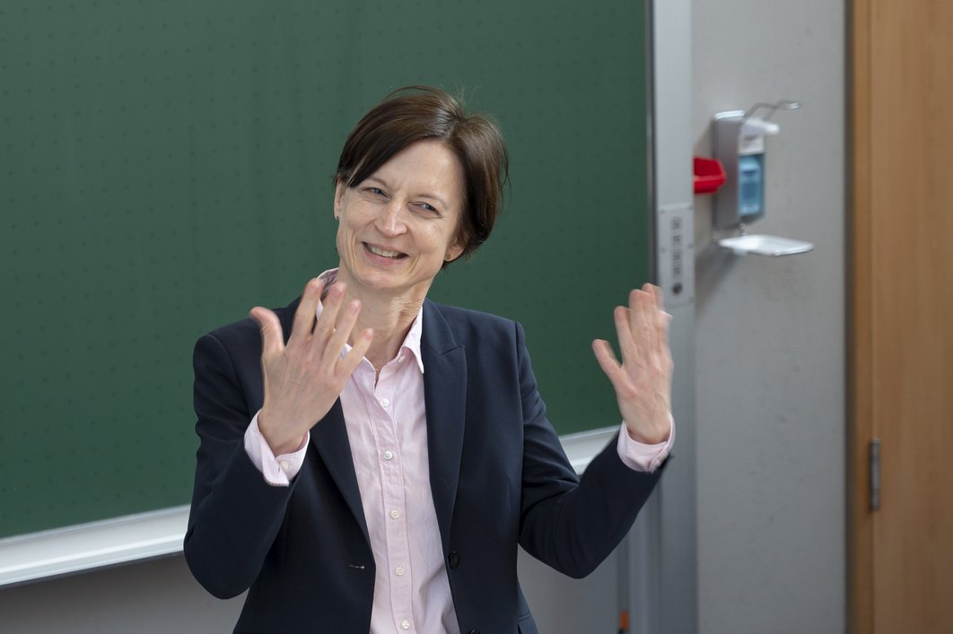 Prof. Simone Hettmer steht in einem Hörsaal und berichtet. Dabei gestikuliert sie und lächelt leicht. Im Hintergrund ist unscharf eine grüne Tafel zu erkennen.