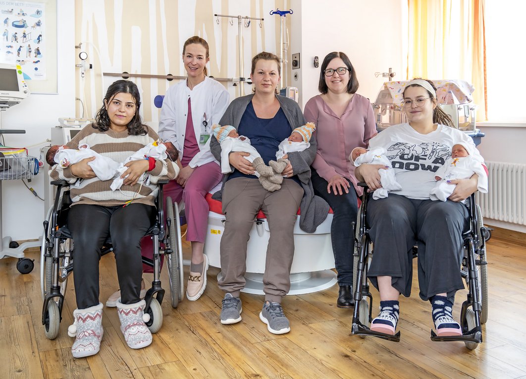 Gruppenfoto: Drei Mütter halten jeweils zwei Babys in den Armen. Zwei von ihnen sitzen im Rollstuhl, eine auf einem Kreißsaal-Bett. Zwischen den Frauen sitzen zwei Frauen in medizinischer Arbeitskleidung.