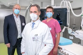 Drei Männer vor einem Herzkatheter-Messplatz. Es ist ein Monitor im Hintergrund zu sehen