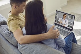 Ein junger Mann sitzt auf einer grauen Couch und hat seinen Arm um die Schultern einer jugen Frau gelegt, die rechts von ihm sitzt. Sie schauen auf den Bildschirm eines Laptops, der vor ihnen auf einem Tisch steht. Auf dem Bilschirm ist eine Ärztin zu sehen.