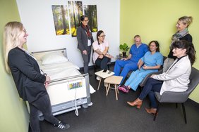 Eine Gruppe von Mitarbeitenden befindet sich im neuen Hybriden Abschiedsraum des Universitätsklinikums Halle (Saale). Einige Personen stehen, einige sitzen. Links an der Wand steht ein Krankenhausbett, rechts ein Sofa und Sessel. An der Wand hängt ein Bild mit Waldmotiv.