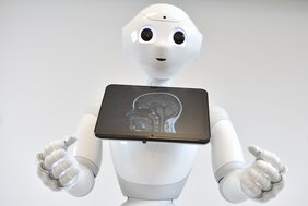 Weißer Roboter in der Größe eines Kindes zeigt auf einem Monitor die Abbildung einer MRT-Aufnahme des Kopfes.